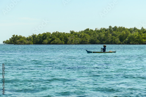 Flores/Indonesia 20290808: A fisherman's boat crossing a calm sea near Maumere, Indonesia There is one man in the boat, wearing straw hat and paddling. There is a small green island in the back. photo