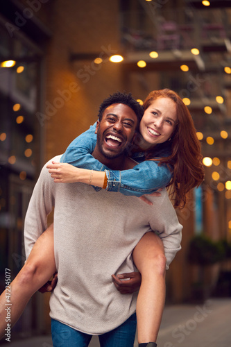 Portrait Of Young Couple Enjoying City Life Heading For Night Out With Man Giving Woman Piggyback