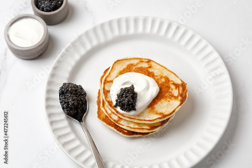 Black Caviar on pancakes with sour cream Holiday morning Appetizer on white plate and marble background with spoon of caviar