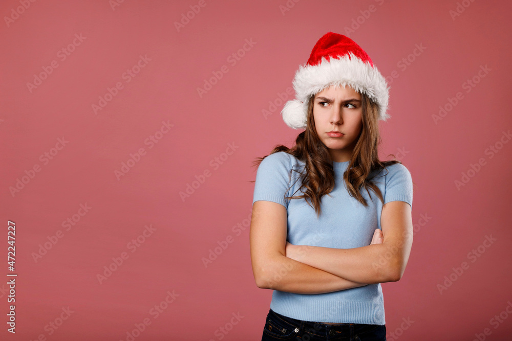 Angry unhappy caucasian girl teenager standing in santa claus red hat looking aside over pink background. Studio portrait. Anti Christmas and New year Holiday concept with copy space. Angry unhappy caucasian girl teenager standing in santa claus red hat looking aside over pink background. Studio portrait. Anti Christmas and New year Holiday concept with copy space.