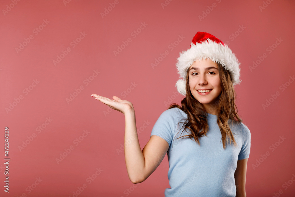 Young happy woman in Santa Claus hat standing over pink background holding your product presenting it. Copy Place for your text. Christmas and New year concept. Young happy woman in Santa Claus hat standing over pink background holding your product presenting it. Copy Place for your text. Christmas and New year concept.
