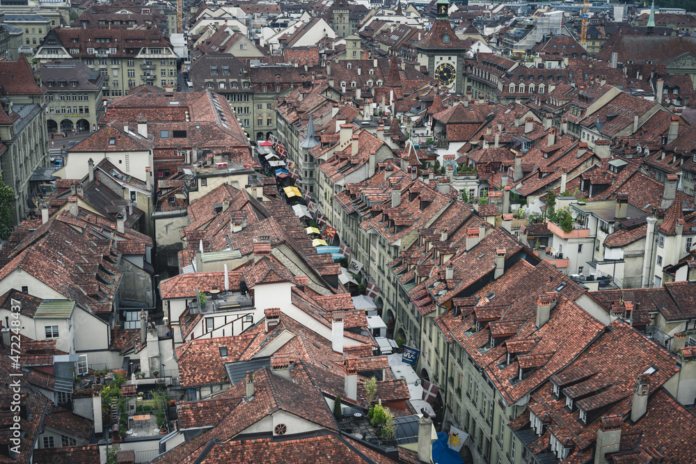 A view of the cityscape of Bern, Switzerland with stone rooftops, streets below, and the farmer's market - a cloudy spring day with green trees and hills in the background