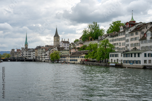 The Limmat River flows next to the cityscape of Zurich, Switzerland on a cloudy spring afternoon - trees and buildings line the river