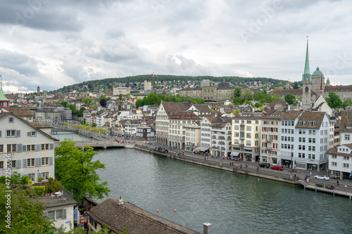 The Limmat River flows next to the cityscape of Zurich, Switzerland on a cloudy spring afternoon - trees and buildings line the river