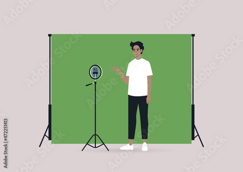 A young male Caucasian blogger recording a video with a tripod, a mobile phone, and a chroma key screen
