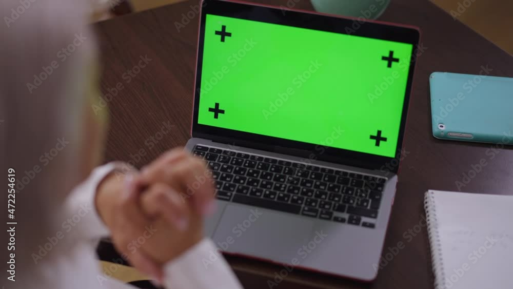 Close-up chromakey laptop on table in office with blurred mature businesswoman sitting indoors. Shooting over shoulder of middle aged Caucasian businesswoman thinking analyzing e-market. Mockup Close-up chromakey laptop on table in office with blurred mature businesswoman sitting indoors. Shooting over shoulder of middle aged Caucasian businesswoman thinking analyzing e-market. Mockup