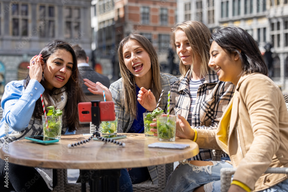 Antwerp, Belgium, May 21, 2021, four mixed race female tourists or students sitting outside in the old city center at a cafe terrace holding a video call using mobile phone. High quality photo
