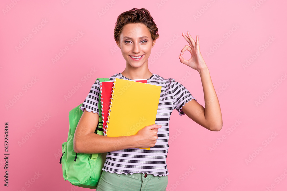 Photo of cool millennial short hairdo student lady hold books show okey wear bag white t-shirt isolated on pink color background Photo of cool millennial short hairdo student lady hold books show okey wear bag white t-shirt isolated on pink color background