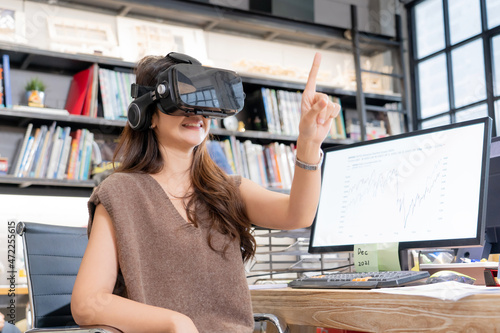 Happy Asian office lady wearing a virtual reality headset and make a gesture touching something in the air while sitting at desk with computer showing a statistic graphs on screen in a blurred office.
