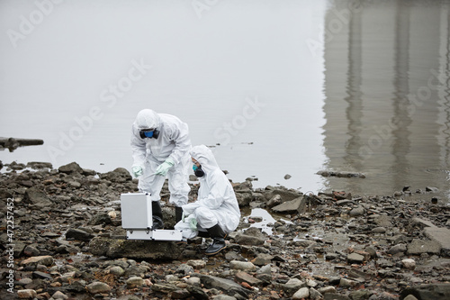 Two workers wearing hazmat suits collecting probe samples by water, toxic waste concept, copy space