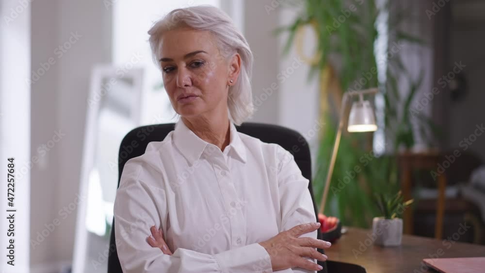 Confident proud mature woman crossing hands in slow motion looking at camera sitting in office indoors. Portrait of attractive slim Caucasian CEO posing at workplace with serious facial expression Confident proud mature woman crossing hands in slow motion looking at camera sitting in office indoors. Portrait of attractive slim Caucasian CEO posing at workplace with serious facial expression