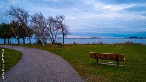 The curving footpath and an empty bench in the park with bare trees. Moody cloudscape at dusk. Twilight seascape over the green at Nut island Pier Park in Quincy, Massachusetts. photo