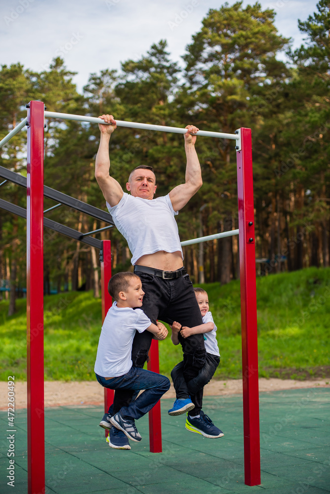Fototapeta premium Caucasian man and two boys doing exercises outdoors. The father pulls himself up on the horizontal bar with his sons on the playground.