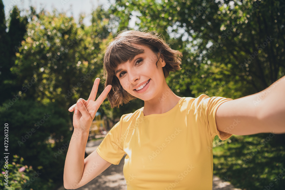 Photo of pretty adorable young woman wear yellow t-shirt smiling recording video showing v-sign outside city street Photo of pretty adorable young woman wear yellow t-shirt smiling recording video showing v-sign outside city street