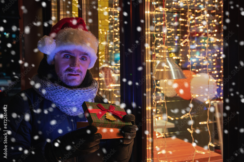 Man in Santas hat with gifts box near illuminated cafe window. Man in Santas hat with gifts box near illuminated cafe window.