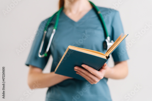 Close-up cropped shot of unrecognizable female doctor intern in uniform with stethoscope reading medical book, standing on white isolated background in studio.
