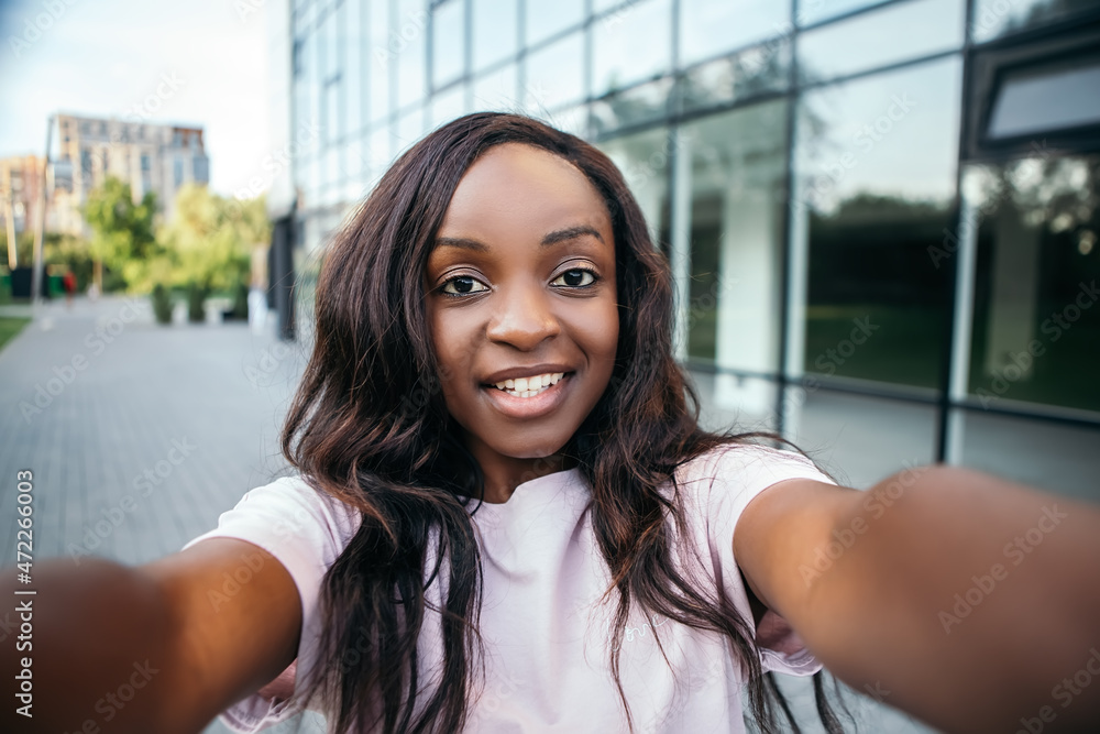 Smiling white teeth beautiful african american pretty woman hold hand phone chatting friends colleague relatives taking selfie send message videochat standing street blur bokeh background