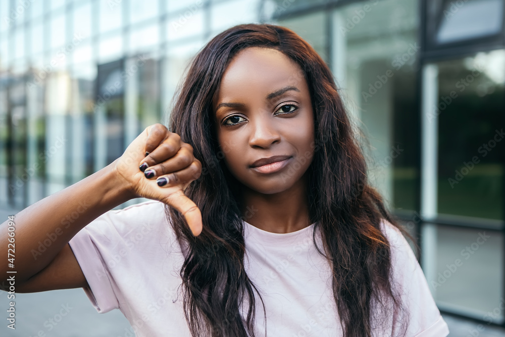 Close up portrait beautiful gesturing african american pretty woman showing thumb down dislike unhappy with dark hair standing outside street dressed t-shirt summer blur bokeh background Close up portrait beautiful gesturing african american pretty woman showing thumb down dislike unhappy with dark hair standing outside street dressed t-shirt summer blur bokeh background