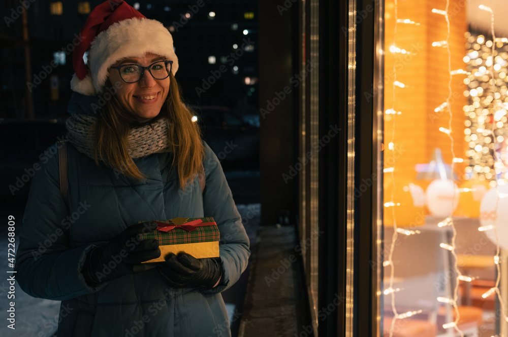 Fototapeta premium Pretty Woman in Santas hat with gifts box near illuminated cafe window. She waves her hand and waits for her beloved partner. Xmas presents holidays, or shopping on New Year or Christmas concept