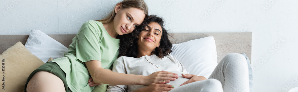 happy woman touching belly of joyful pregnant girlfriend in bedroom, banner happy woman touching belly of joyful pregnant girlfriend in bedroom, banner