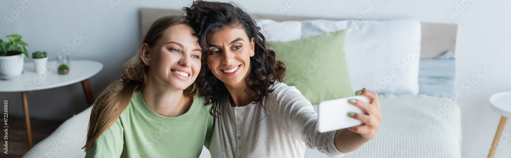 curly woman taking selfie with happy girlfriend in bedroom, banner curly woman taking selfie with happy girlfriend in bedroom, banner
