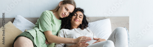 happy woman touching belly of joyful pregnant girlfriend in bedroom  banner