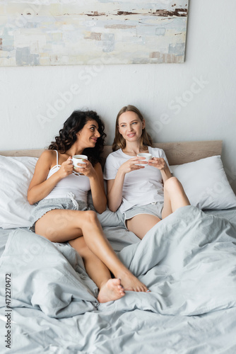 pleased young lesbian couple holding cups of coffee in bed