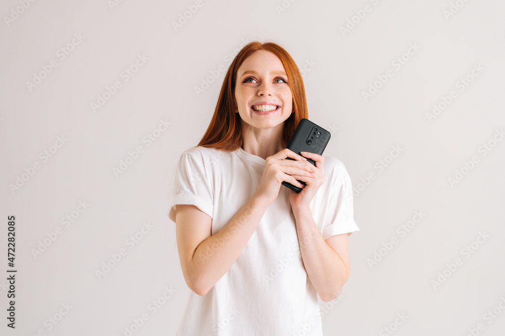 Studio portrait of happy dreaming young woman with wide smile reading good online message using mobile phone on white isolated background. Cheerful lady holding smartphone in hands and looking up. Studio portrait of happy dreaming young woman with wide smile reading good online message using mobile phone on white isolated background. Cheerful lady holding smartphone in hands and looking up.