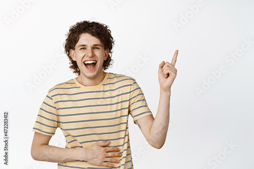 Carefree young man laughing at smth funny, pointing finger at upper right corner and smiling, white background