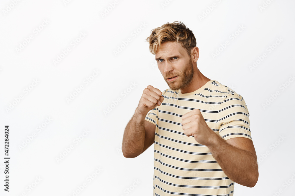 Strong handsome blond man standing in boxing fighter pose, clenching fists ready for fight, posing in casual t-shirt over white background Strong handsome blond man standing in boxing fighter pose, clenching fists ready for fight, posing in casual t-shirt over white background