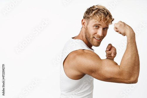 Portrait of handsome fitness athlete flexing biceps, showing strong muscle arm and smiling, workout in gym, standing over white background