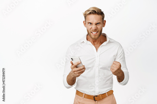 Angry blond man holding cellphone, clenching fist furious and upset, standing over white background