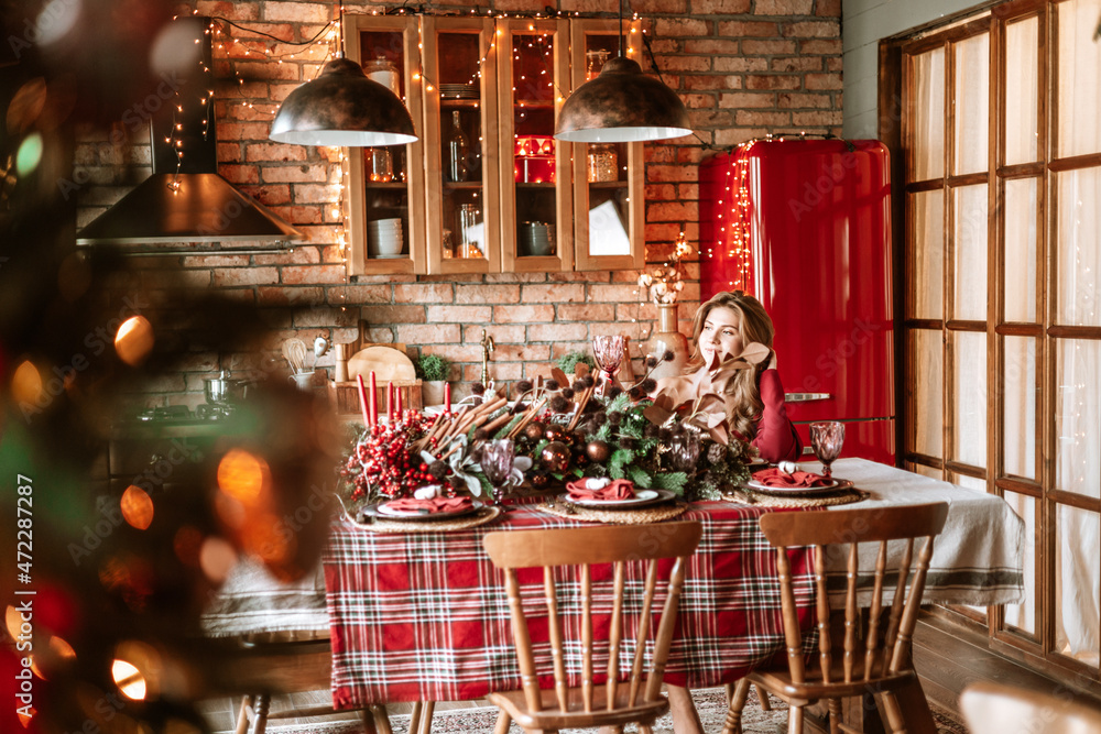 beautiful young woman in red dress with glass of wine stands near Christmas tree in living room decorated for celebration of Christmas and New Year in stylish interior, table set for family dinner