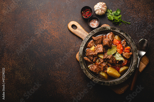 Delicious beef meat stew dish with potatoes, carrot and gravy in rustic metal bowl with spoon, bunch of fresh parsley, garlic cloves, spices on brown concrete background top view space for text