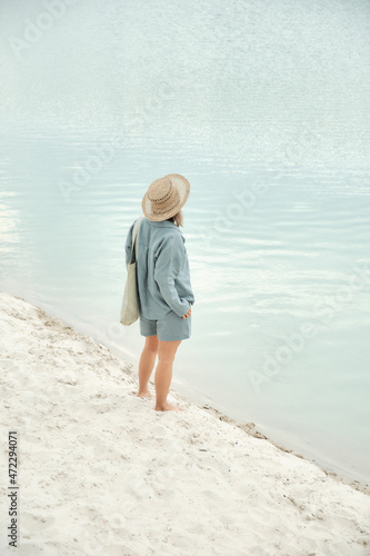 Young girl in natural linen clothes and a straw hat, barefoot on a white sand beach, holding a linen bag. Back view.