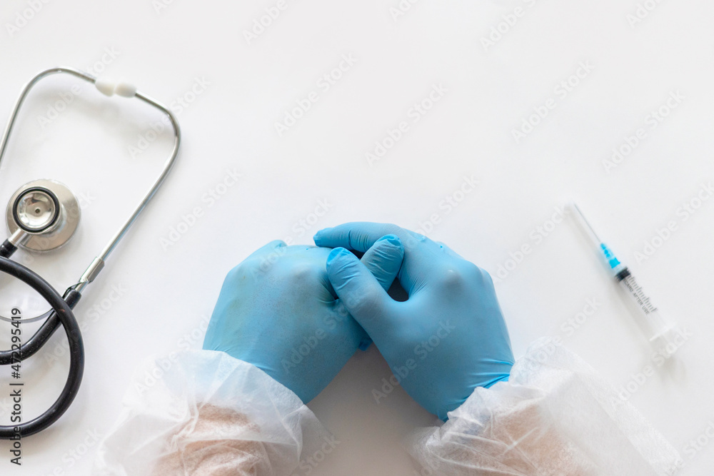Fototapeta premium hands of young doctor in protective suit and blue protective gloves on table next to stethoscope and syringe for injecting vaccine