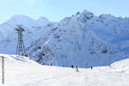 A wide ski slope against a backdrop of sharp snowy mountains and blue skies. There is a cable car on the side of the slope. On the slope of snowboarders and skiers. © VeNN