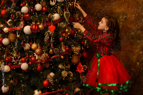 Beautiful little girl in a Christmas shirt and red skirt is decorating a Christmas tree.Christmas mood