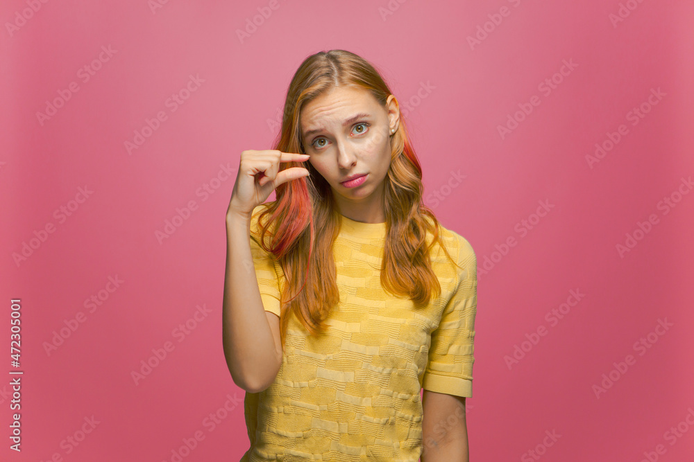 Disappointed girl showing small size with pity on pink studio background. Young woman showing measurement with fingers Disappointed girl showing small size with pity on pink studio background. Young woman showing measurement with fingers