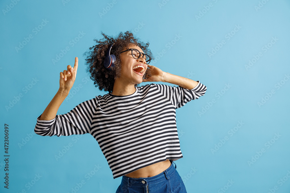 Cheerful multiracial lady enjoying favorite songs and laughing while moving to the rhythm of music. Isolated on blue background Cheerful multiracial lady enjoying favorite songs and laughing while moving to the rhythm of music. Isolated on blue background