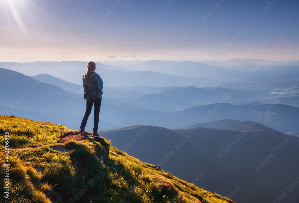 Girl with backpack on mountain peak with green grass looking at beautiful mountain valley in fog at sunset in summer. Landscape with sporty young woman, foggy hills, sky. Travel and tourism. Hiking