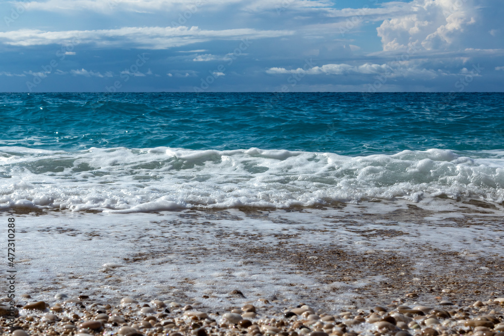 Scenic shining white foam on pebble beach with blue stormy waves and epic cloudscape on coast of Lefkada island in Greece. Summer nature travel to Ionian Sea