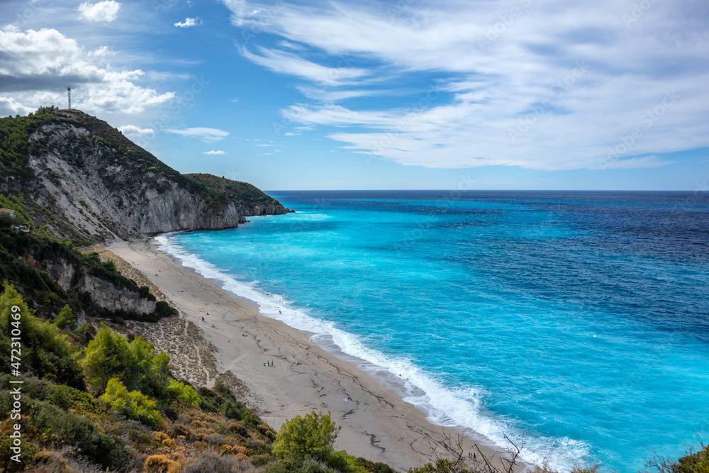 Azure vibrant waves on green rocky steep coast of Lefkada island. Mylos sandy beach with epic cloudscape in Greece. Summer nature vacation travel to Ionian Sea