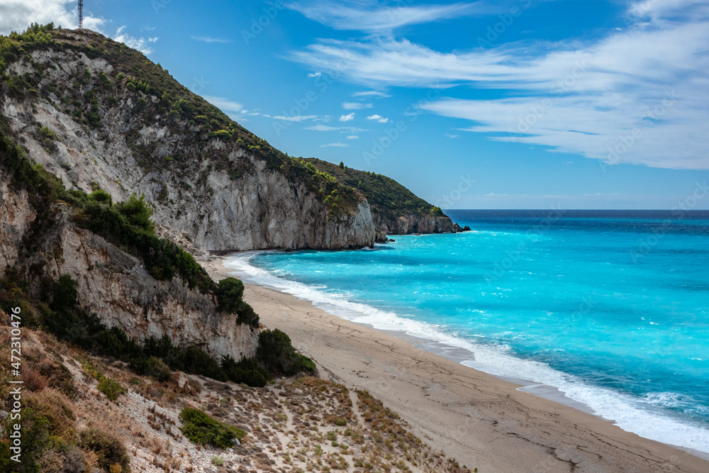 Steep rocky shore of Mylos sandy beach with azure vibrant sunny waves and blue sky. Coast of Lefkada island in Greece. Summer vacation travel to Ionian Sea