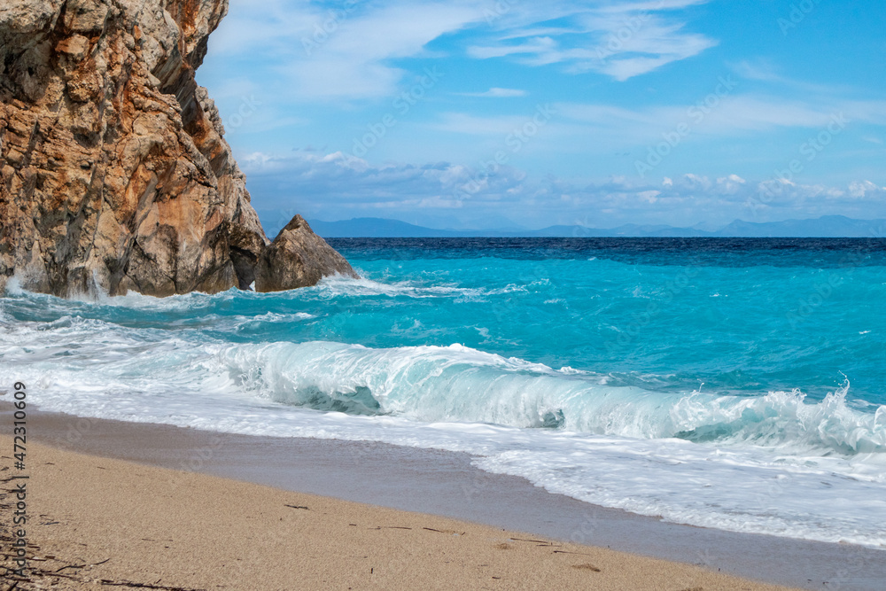 Azure sunny stormy waves with white foam breaking rocky coast. Sandy beach with blue sky on Lefkada island in Greece. Summer nature travel to Ionian Sea