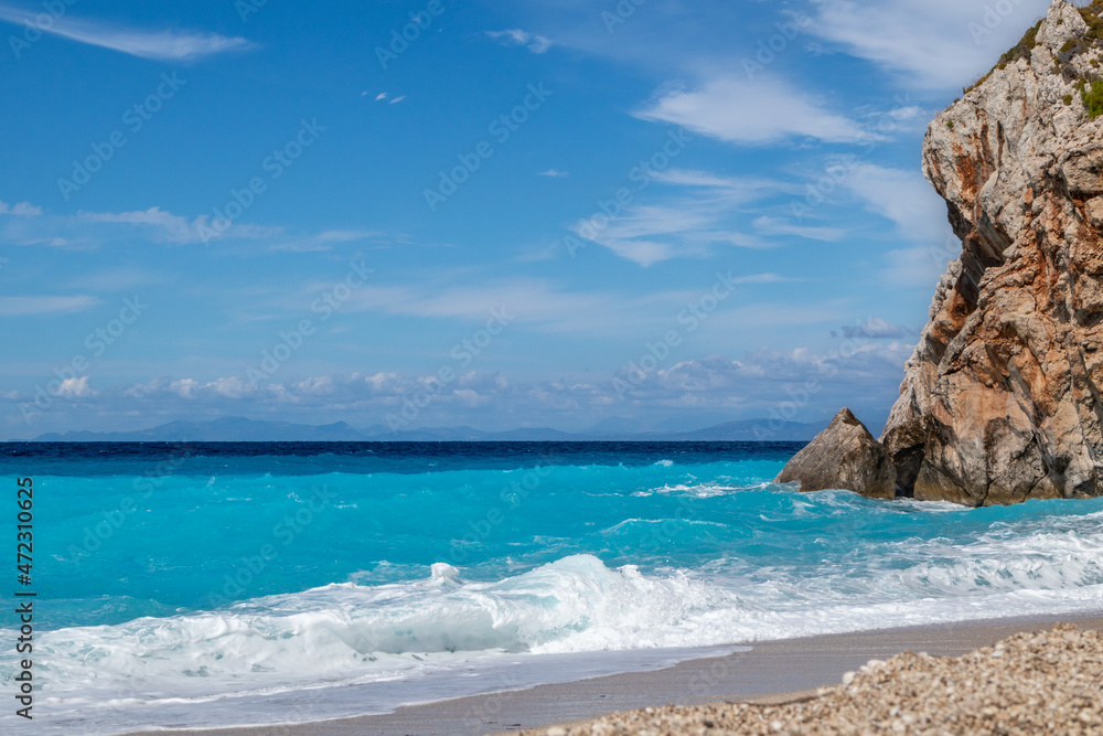 Blue vibrant waves with white foam breaking rocky coast. Mylos sandy beach with scenic sky on Lefkada island in Greece. Summer nature travel to Ionian Sea