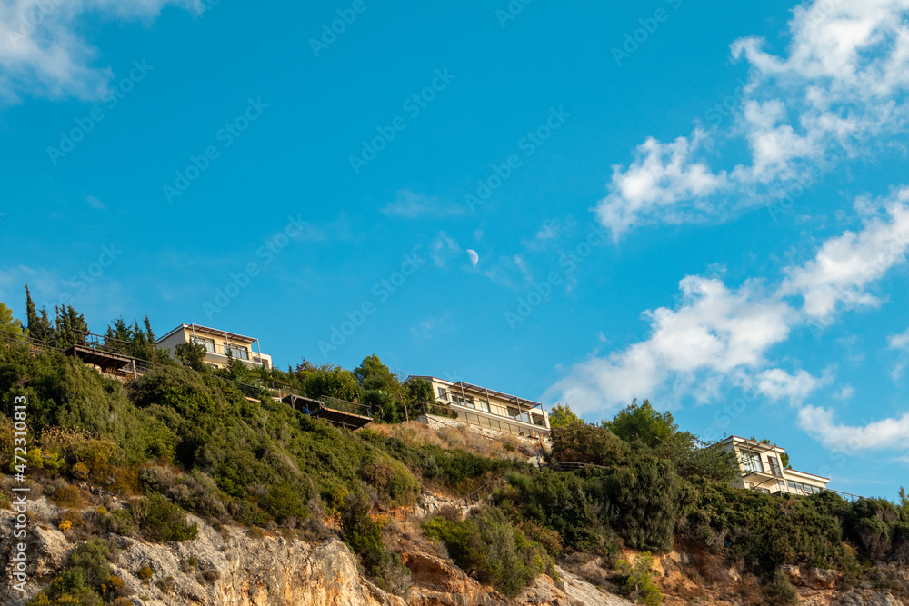 Scenic vibrant landscape with Greek recreation villa houses on green high hills on blue scenic sky with clouds and young moon on Lefkada island, Greece. Travel in summer