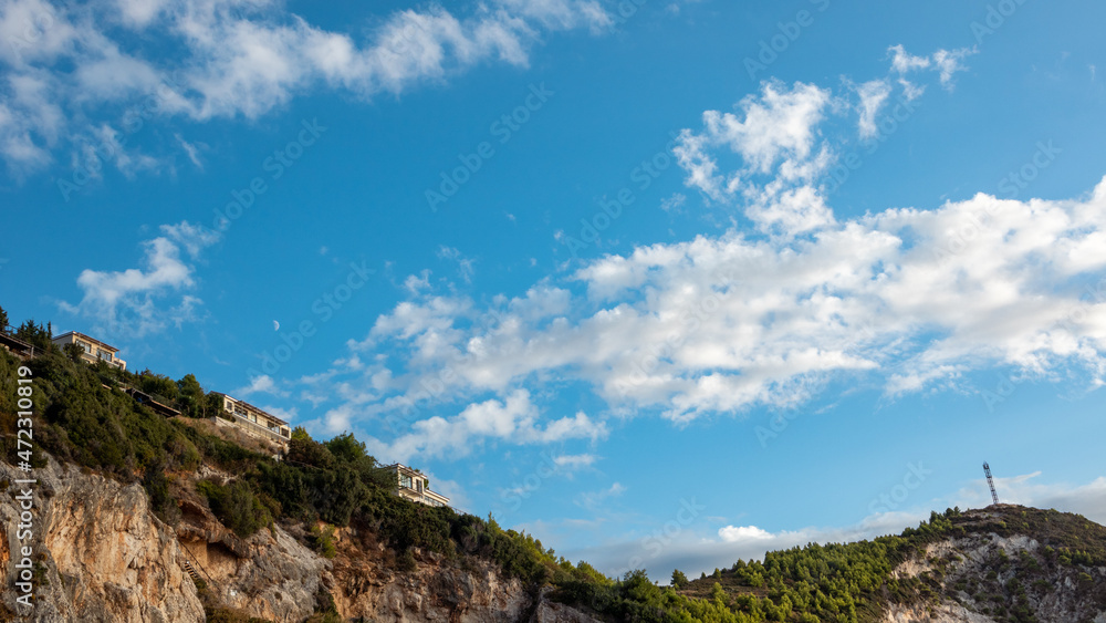 Skyscape view on Greek recreation villa houses on green high hills. Blue scenic sky with clouds and young moon on Lefkada island, Greece. Travel in summer Skyscape view on Greek recreation villa houses on green high hills. Blue scenic sky with clouds and young moon on Lefkada island, Greece. Travel in summer
