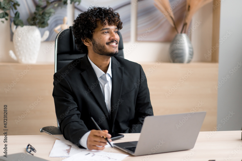 Indian positive businessman, in formal clothes, sitting at the table in office, while working in a laptop, looking positively aside, thinking about a startup or dreaming about a future vacation Indian positive businessman, in formal clothes, sitting at the table in office, while working in a laptop, looking positively aside, thinking about a startup or dreaming about a future vacation