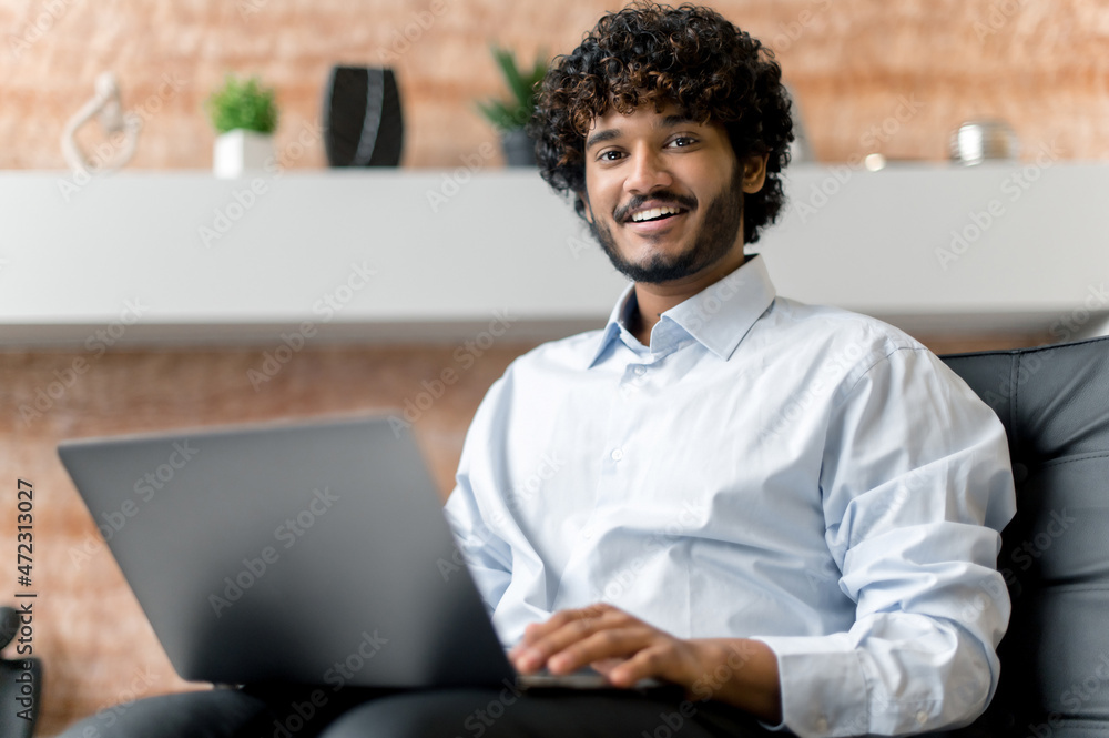 Indian positive confident curly-haired young adult guy, freelancer or manager in shirt, sitting on armchair with laptop looking at the camera and smiling friendly Indian positive confident curly-haired young adult guy, freelancer or manager in shirt, sitting on armchair with laptop looking at the camera and smiling friendly